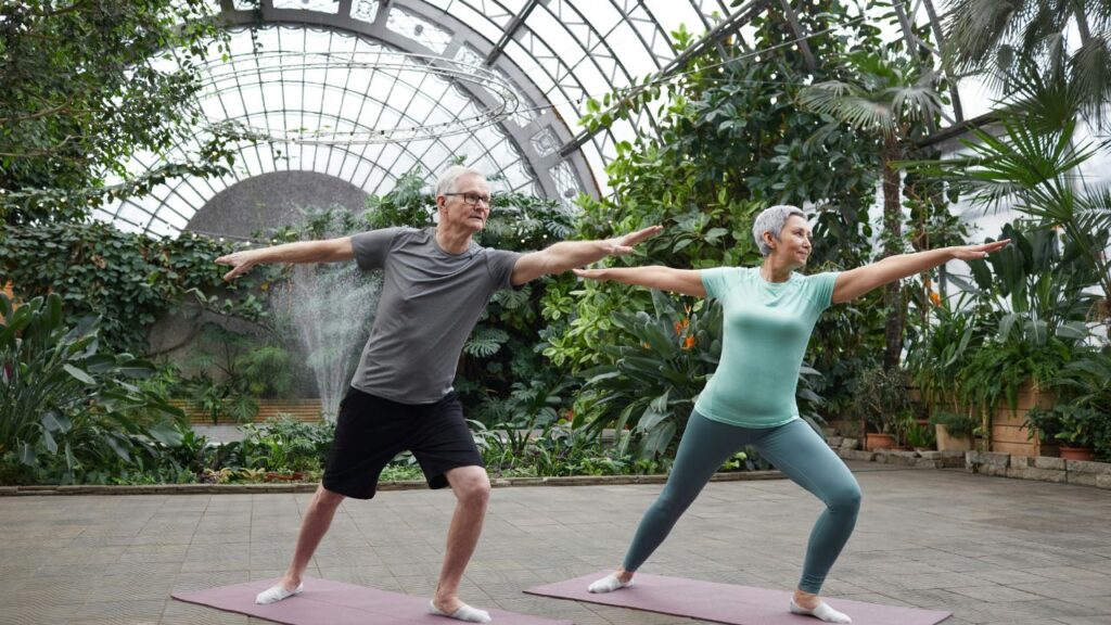 Dos personas mayores activas (hombre y mujer) haciendo yoga o estiramientos en un entorno natural y luminoso, representando la salud y bienestar en el Senior Living.
